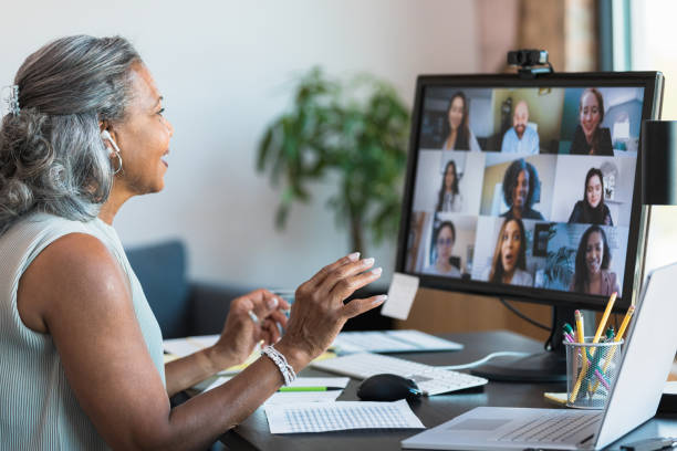 The senior adult woman gestures and smiles as she leads a meeting with a multi-ethnic group of co-workers.